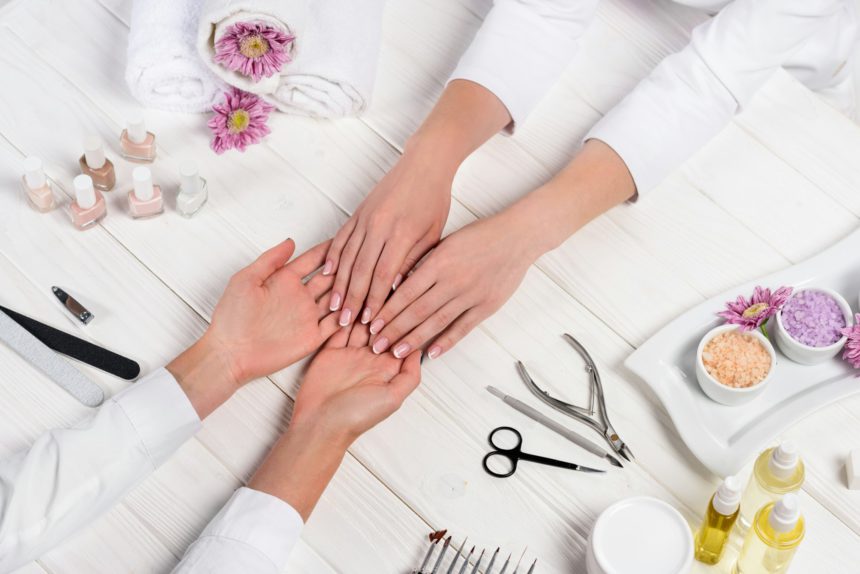 cropped image of manicurist looking at hands of woman at table with flowers, towels, nail polishes,