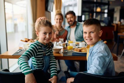 Happy children with parents having a breakfast at hotel restaurant and looking at camera.